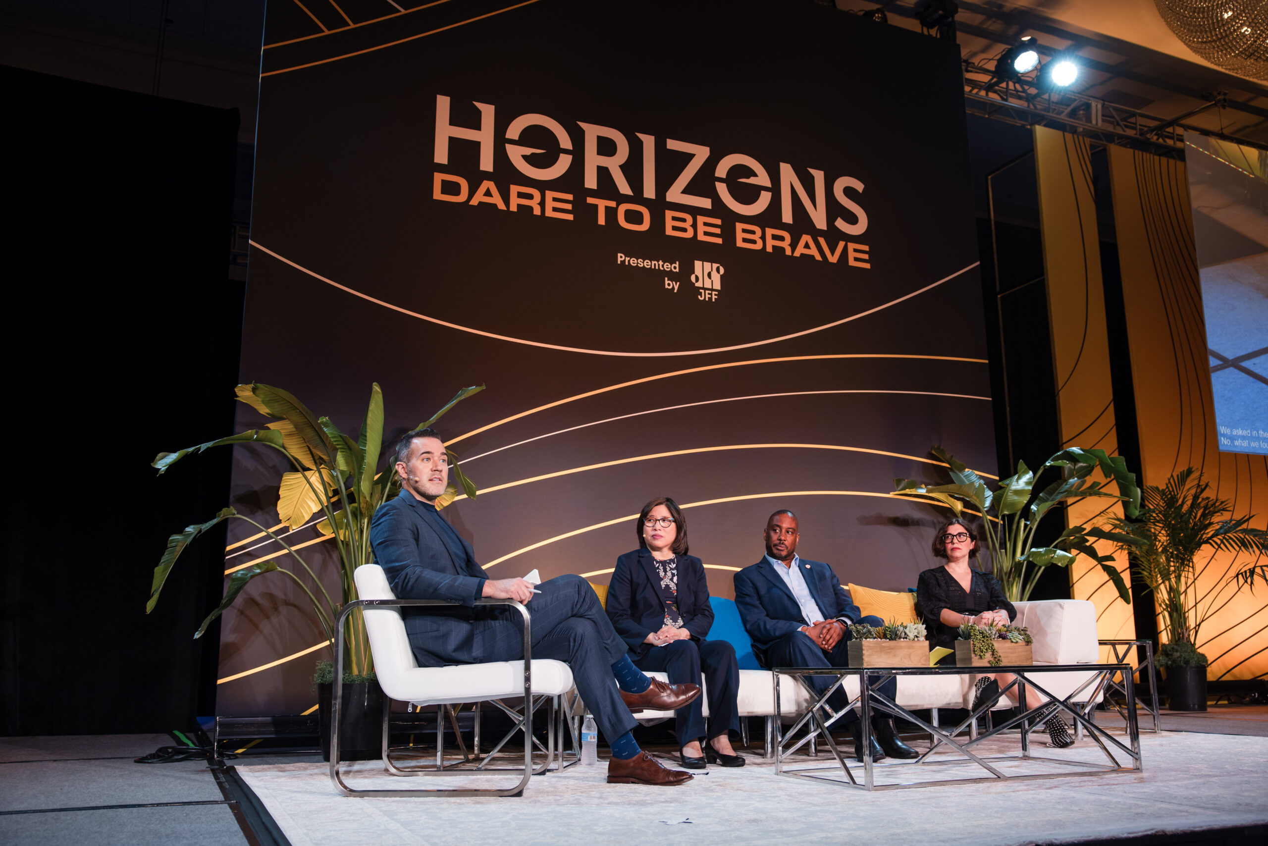 Four people sit on a stage in front of a large "Horizons: Dare to be Brave" sign, engaged in a panel discussion. Two large plants and a coffee table are also on the stage.