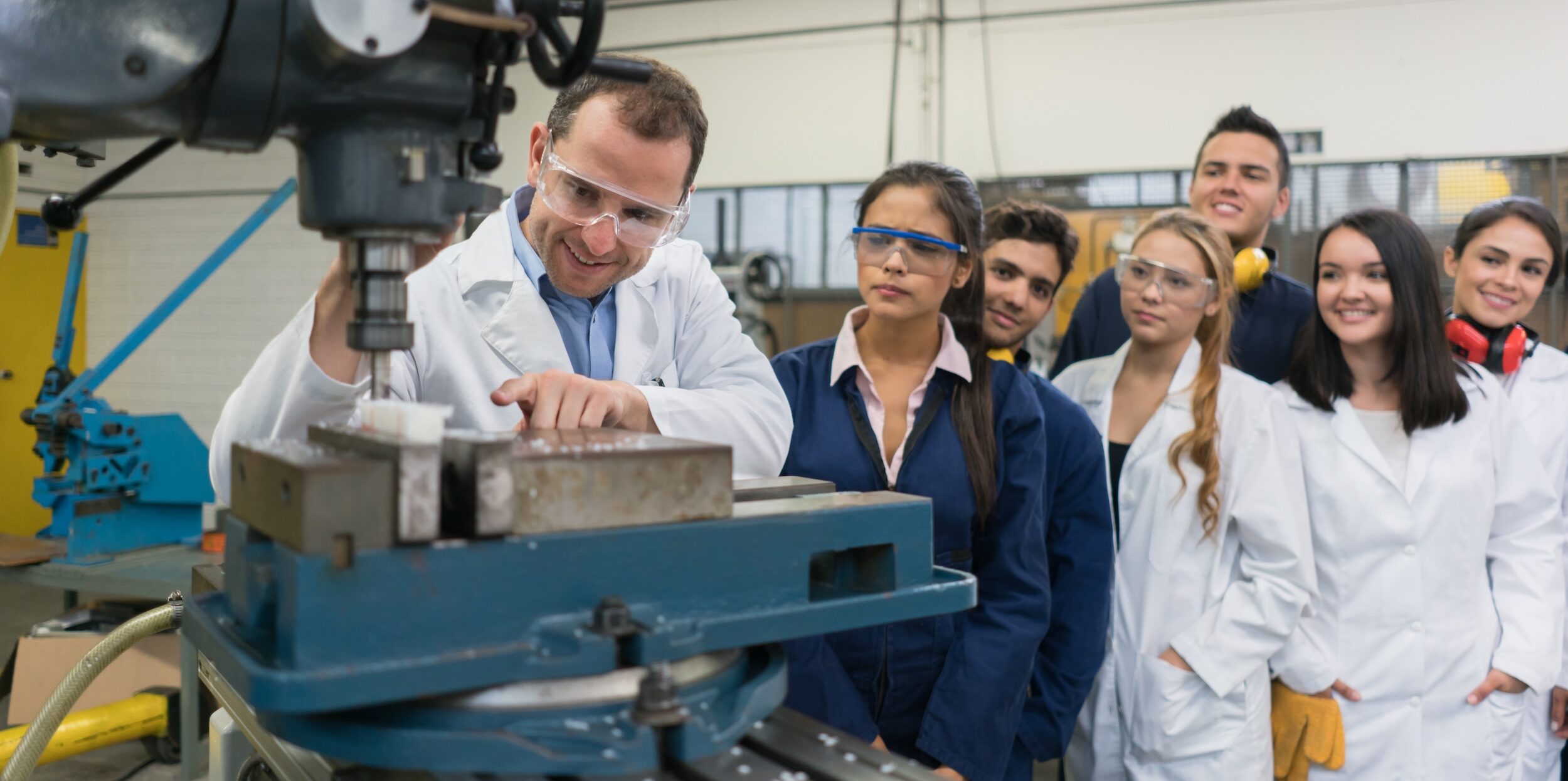 A group of people in lab coats and safety glasses observe a man operating a milling machine in a workshop or laboratory setting.