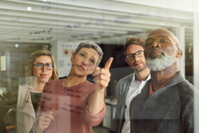 Four people stand in front of a glass wall with notes, discussing and analyzing written ideas in a modern office setting.