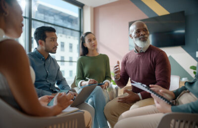 A group of five people sit together in a modern office, engaged in discussion with notepads and a laptop.