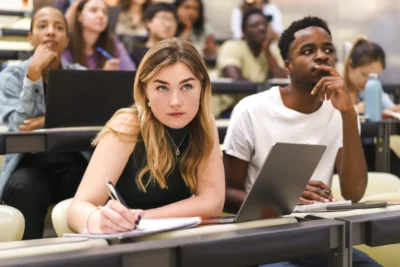 Students sitting in a lecture hall, attentively listening, with notebooks and laptops open in front of them.