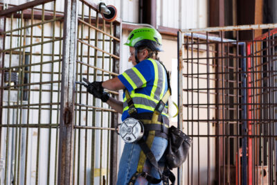 A construction worker in safety gear adjusts a metal gate at a worksite, wearing a helmet, gloves, and a harness.