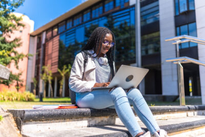 A young woman with headphones sits on outdoor steps, using a laptop, with books beside her and a modern building in the background.