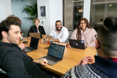 Five people sitting around a conference table with laptops, smiling and having a discussion in a modern office setting.