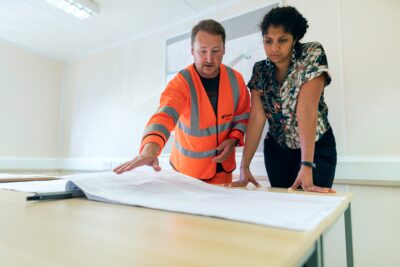Two people stand at a table reviewing architectural plans; one wears a high-visibility jacket and gestures toward the documents.