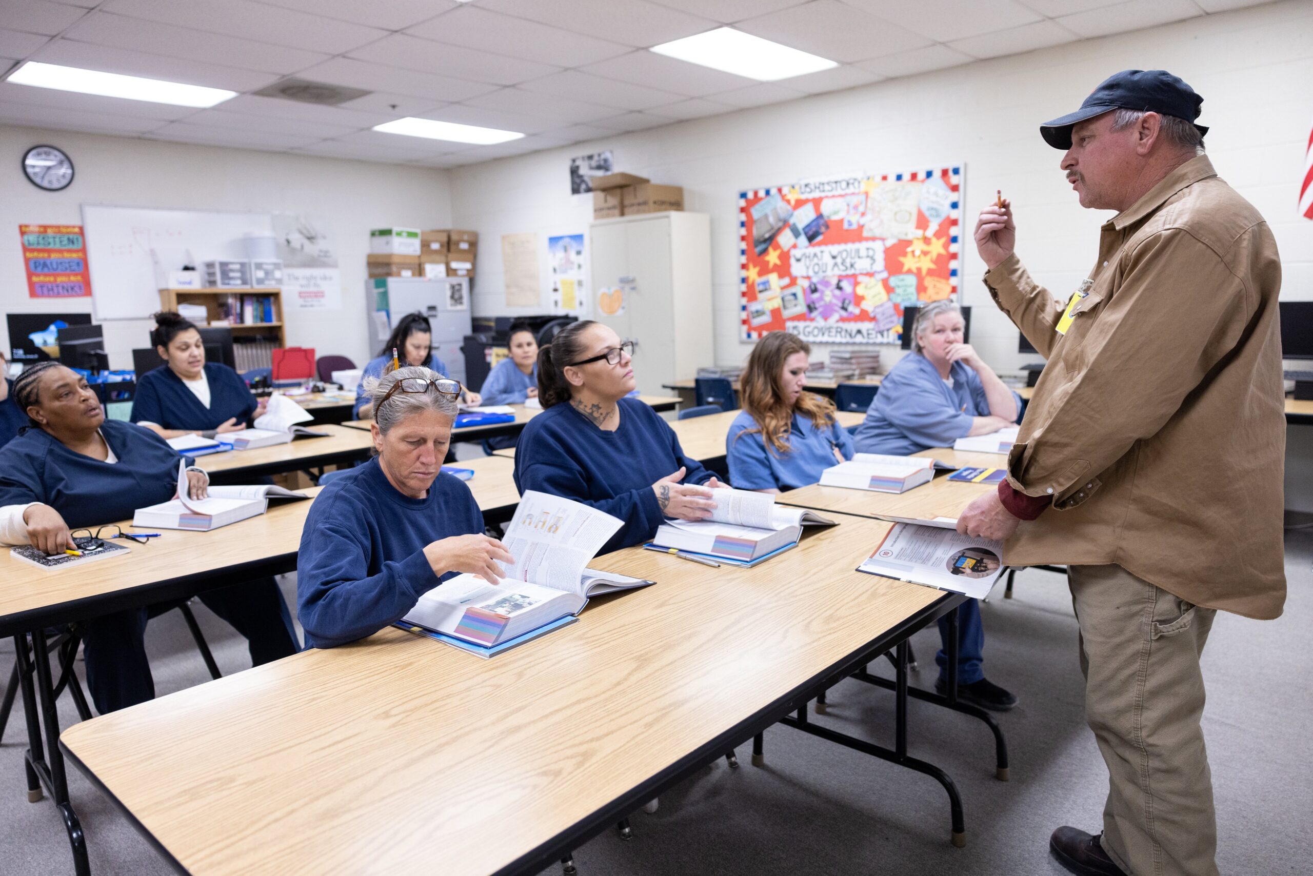 An instructor stands at the front of a classroom speaking to adult students seated at desks with open books in a brightly lit room.
