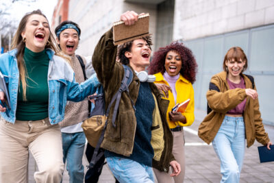 Five young adults walk outside together, laughing and smiling; one person in front raises a notebook in celebration.