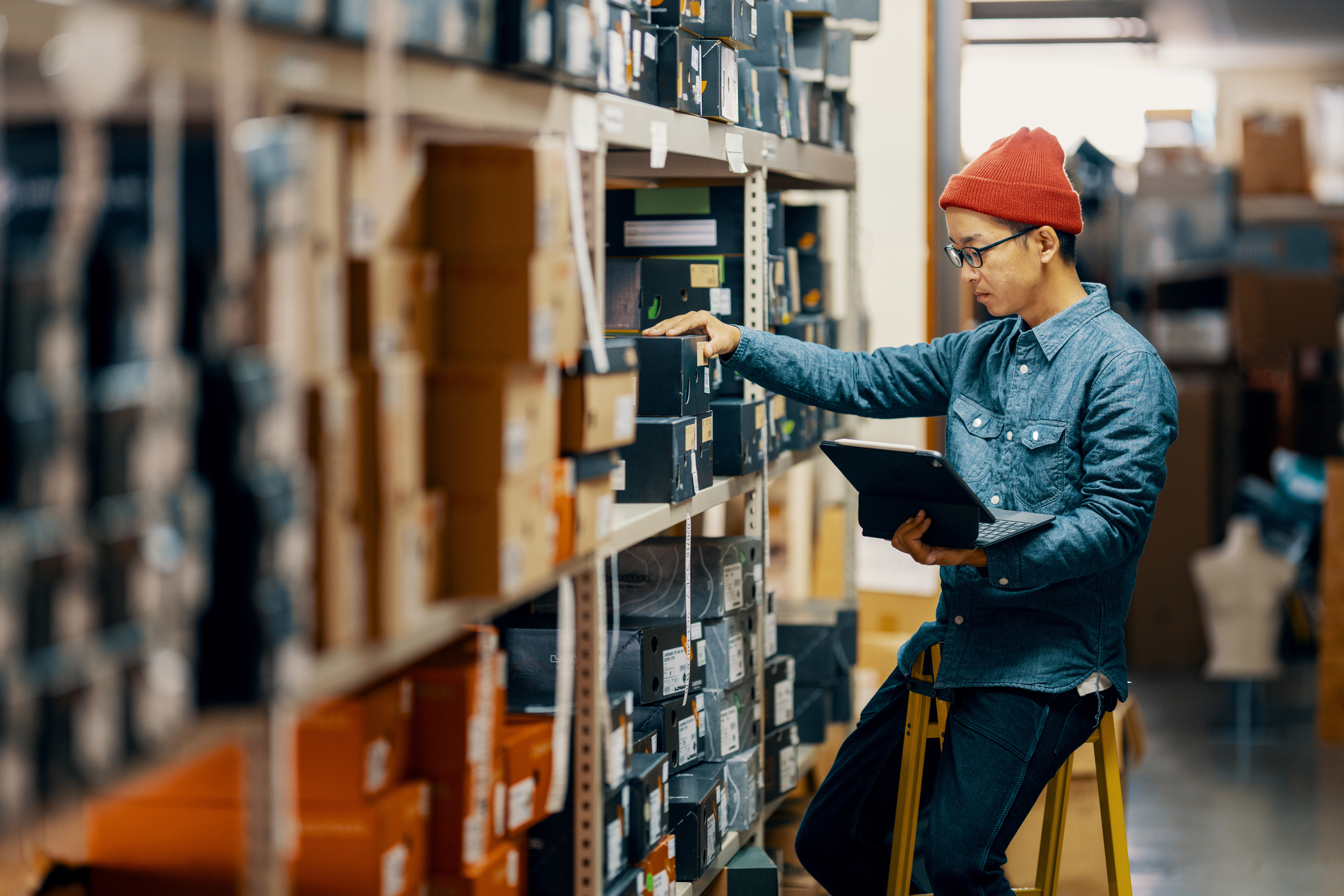A person wearing a red beanie and denim shirt checks inventory with a laptop in a warehouse, surrounded by shelves filled with boxes.