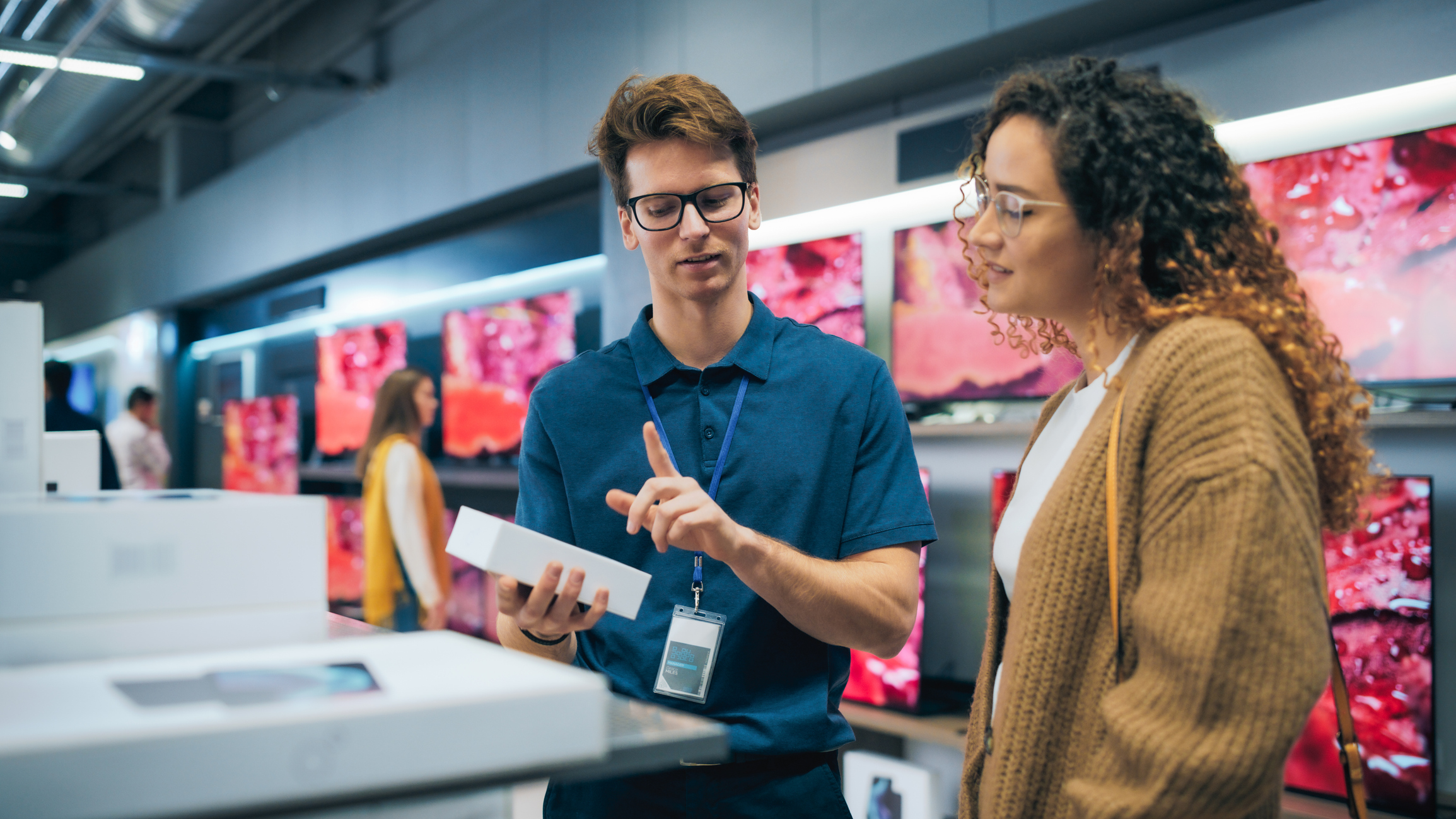A store employee shows a product box to a customer in an electronics store, with televisions displayed on the wall in the background.