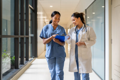 Two healthcare professionals, one in scrubs and one in a lab coat with a stethoscope, review notes on a clipboard while walking down a hallway.