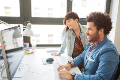 Two people work together at a computer desk in a brightly lit office, smiling as they look at the monitor—collaborating on reimagining postsecondary success in the age of AI.