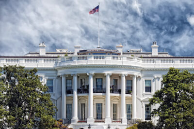 The White House with the American flag flying on top, viewed from the front with columns and greenery in the foreground.