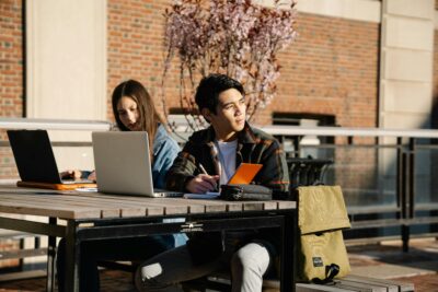 Two students sit at an outdoor table with laptops and notebooks, working individually. A tan backpack rests on the table beside them and a brick building is in the background.