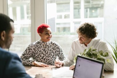Three people sit at a table in a bright office, talking and smiling, with a laptop and documents in front of them.