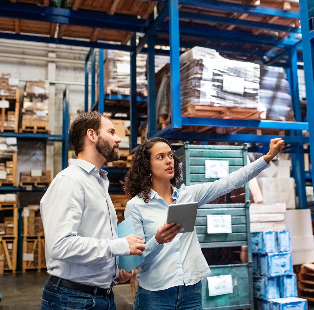 Two people in a warehouse discussing inventory; one holds a tablet while the other gestures toward shelves stacked with boxes and packages.