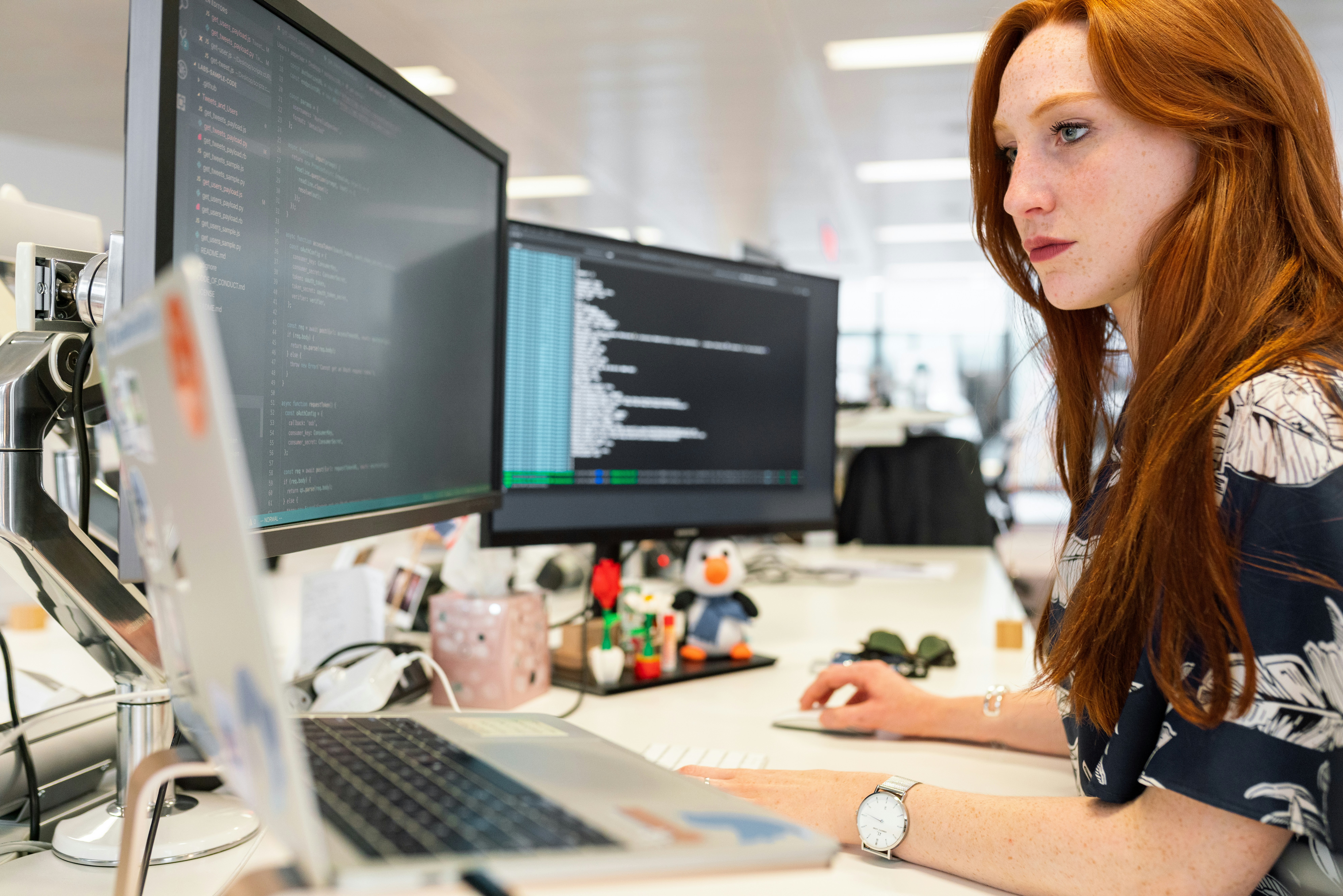 A woman with long red hair works at a desk with multiple monitors displaying code in a modern office setting.