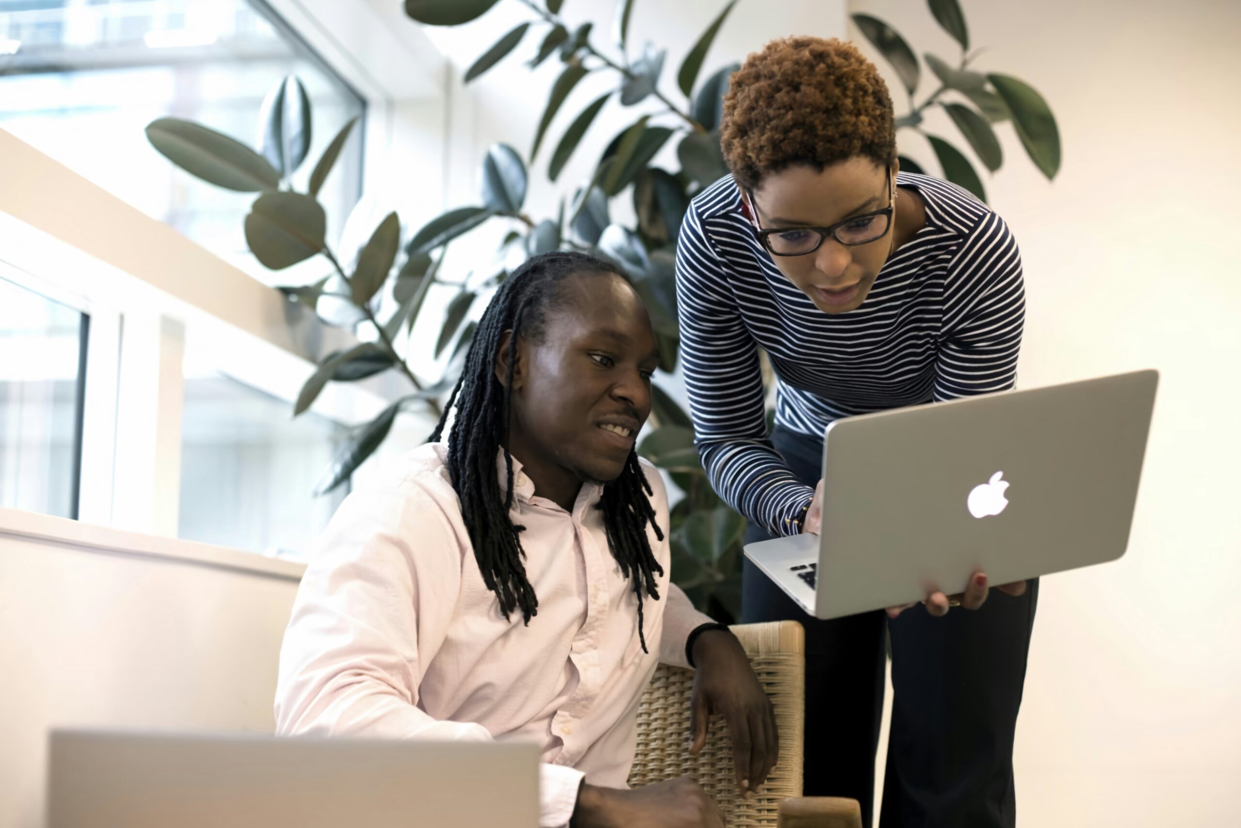 Two people look at a laptop together in an office setting, one sitting and one standing, with a large plant in the background.