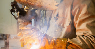 A person wearing protective gloves and a jacket welds metal, producing sparks and smoke.
