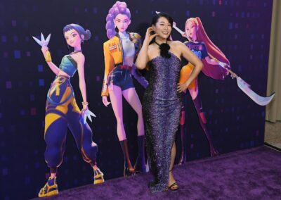 A woman in a sparkly purple dress poses on a purple carpet in front of a backdrop featuring three animated female characters.