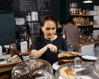 A person slices a piece of layered chocolate cake behind a cafe counter, with cake displays and coffee equipment visible in the background.