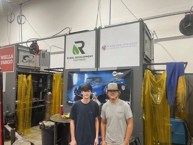 Two young men stand in front of welding booths with signage for Rural Development Partners and Tennessee Colleges of Applied Technology. Workshop equipment and safety curtains are visible.