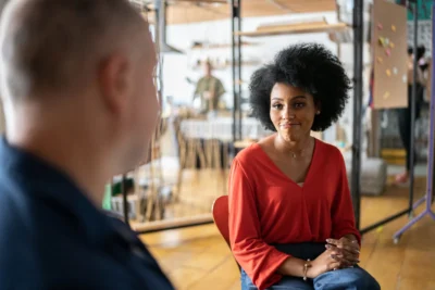 Two people sit indoors having a conversation; the woman in the foreground wears a red top and smiles slightly while listening.