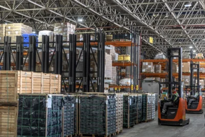 A warehouse interior with shelves stacked with boxes and pallets; a worker operates a forklift among the stored goods.