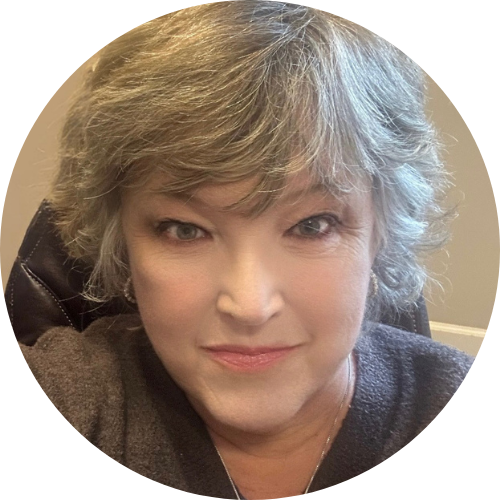 A person with short, gray hair and light skin is sitting indoors, looking at the camera and smiling slightly.