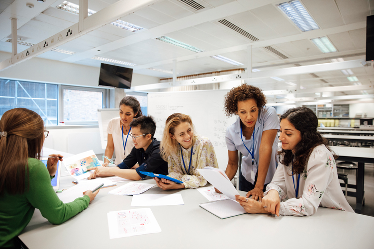 Five people in an office or classroom setting sit and stand around a table, discussing documents and charts, with papers and laptops in front of them.