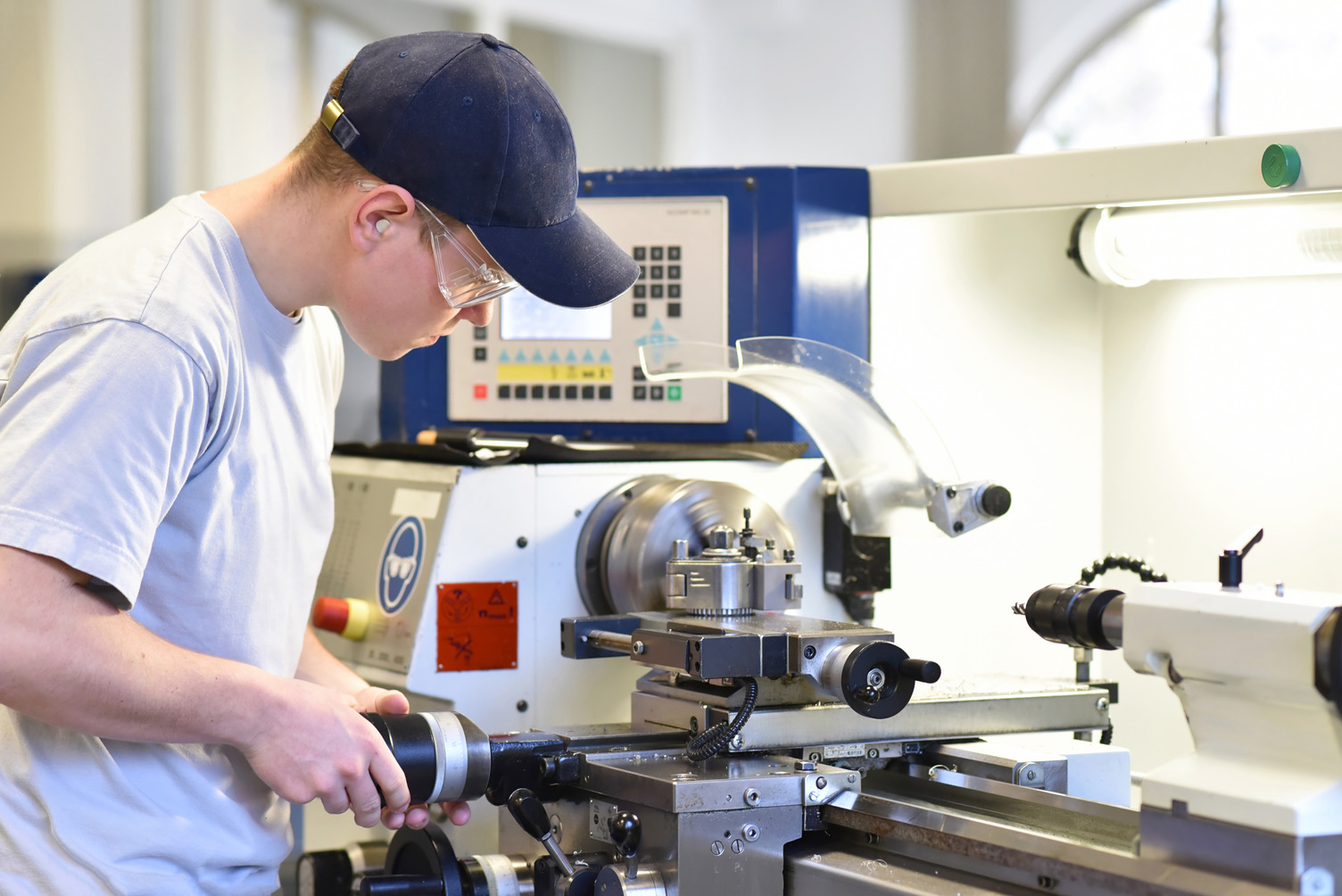 A person wearing safety glasses and a cap operates a metal lathe machine in an industrial workshop setting.