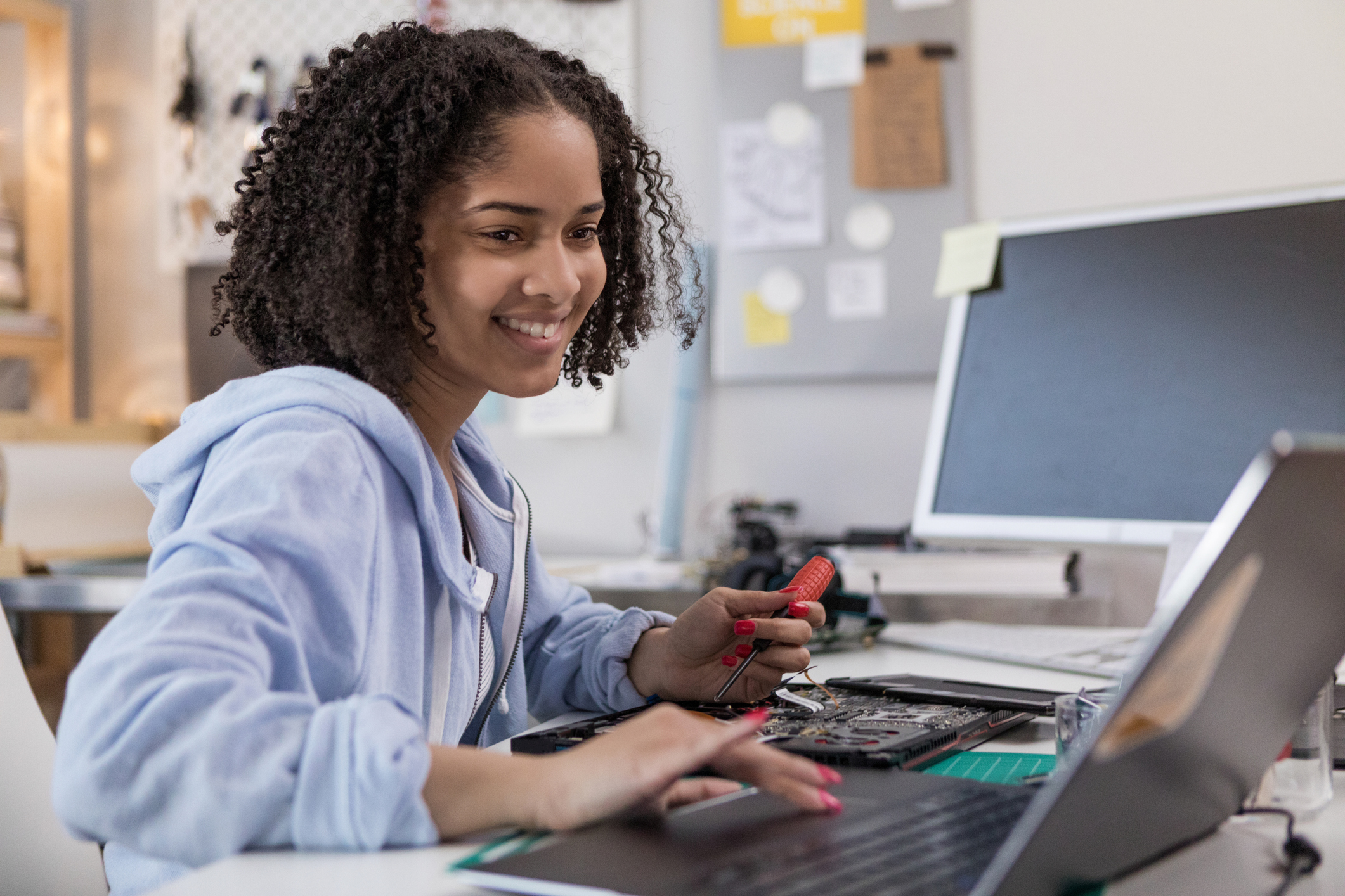 A woman smiles while using a screwdriver to repair or assemble a laptop at a desk, with tools and computer parts in the background.