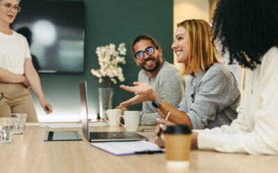 Four people are having a discussion around a conference table, with laptops, notebooks, and coffee cups in front of them. One person is speaking and gesturing with a hand.