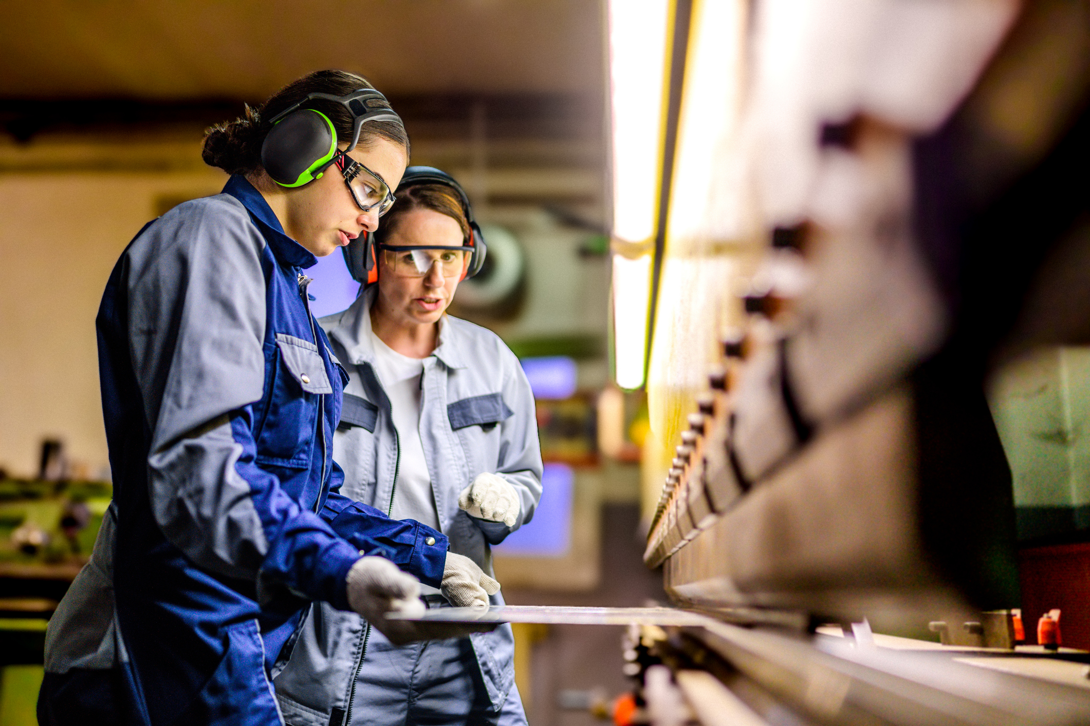 Two people wearing protective gear work together at a metal bending machine in an industrial workshop.