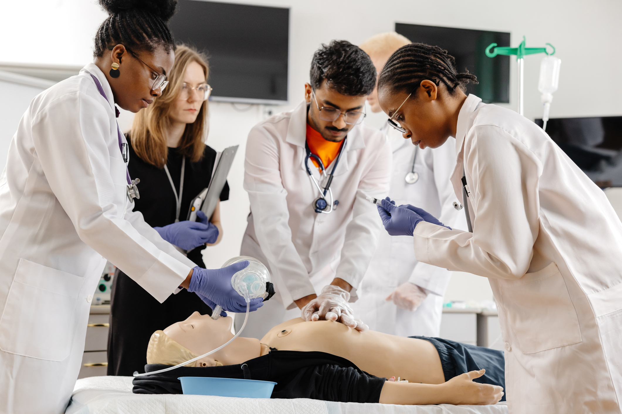 Medical students in lab coats practice emergency procedures on a medical manikin in a clinical training room.