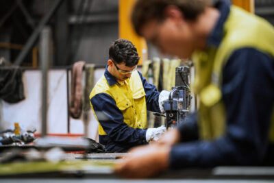 Two workers wearing yellow safety vests operate machinery in an industrial workshop. One man is in focus working with equipment, while another is blurred in the foreground.