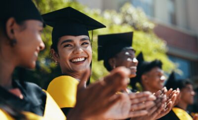 A group of graduates in caps and gowns stand outdoors, smiling and applauding during a graduation ceremony.
