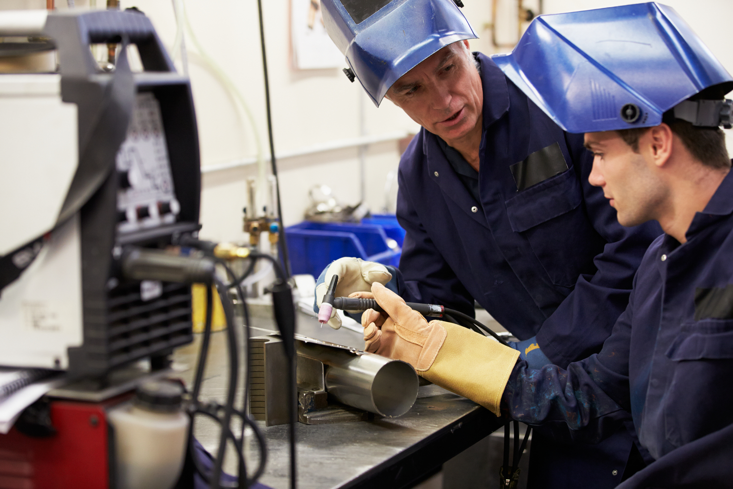 Two workers in protective gear operate welding equipment at a workbench, with one instructing the other. Various tools and machinery are visible in the background.