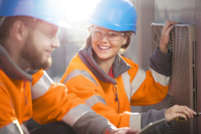Two workers in orange high-visibility jackets and blue helmets smile while servicing an outdoor HVAC unit.