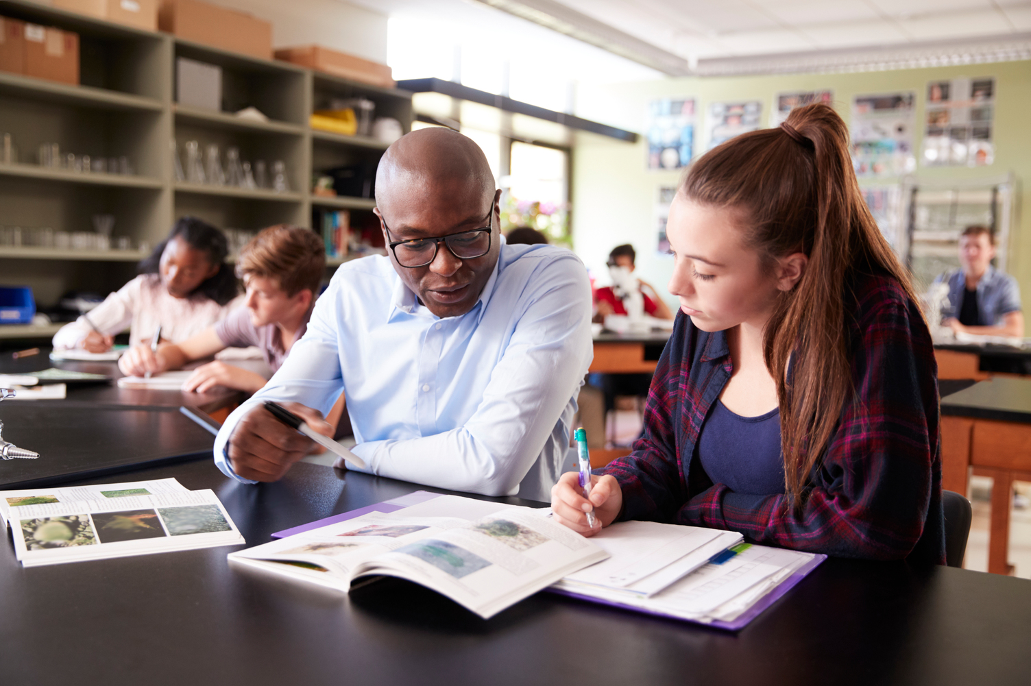 A teacher sits at a science lab table, helping a student with her textbook and notes; other students work in the background.