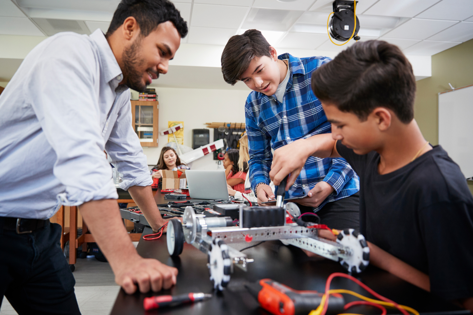 A teacher supervises two students assembling a robotic vehicle at a classroom table, with tools and electronic parts scattered around.