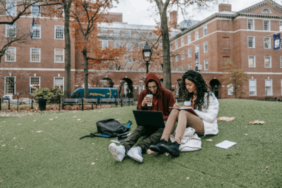 Two students sit on grass with backpacks, using a laptop and tablet while drinking coffee, with campus buildings and autumn trees in the background.