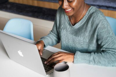 A person in a green sweater is typing on a silver laptop at a desk with a gray mug nearby.