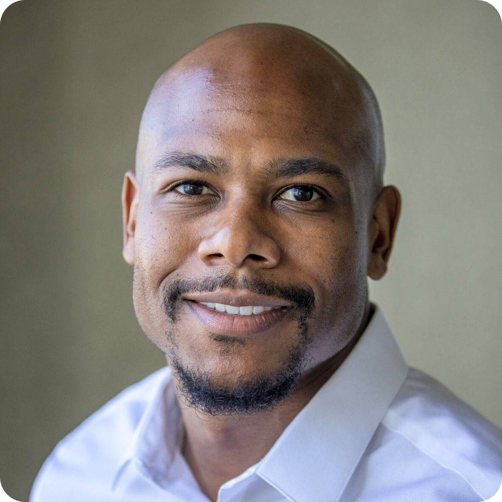 A man with a shaved head and short beard, wearing a white collared shirt, smiles at the camera against a neutral background.