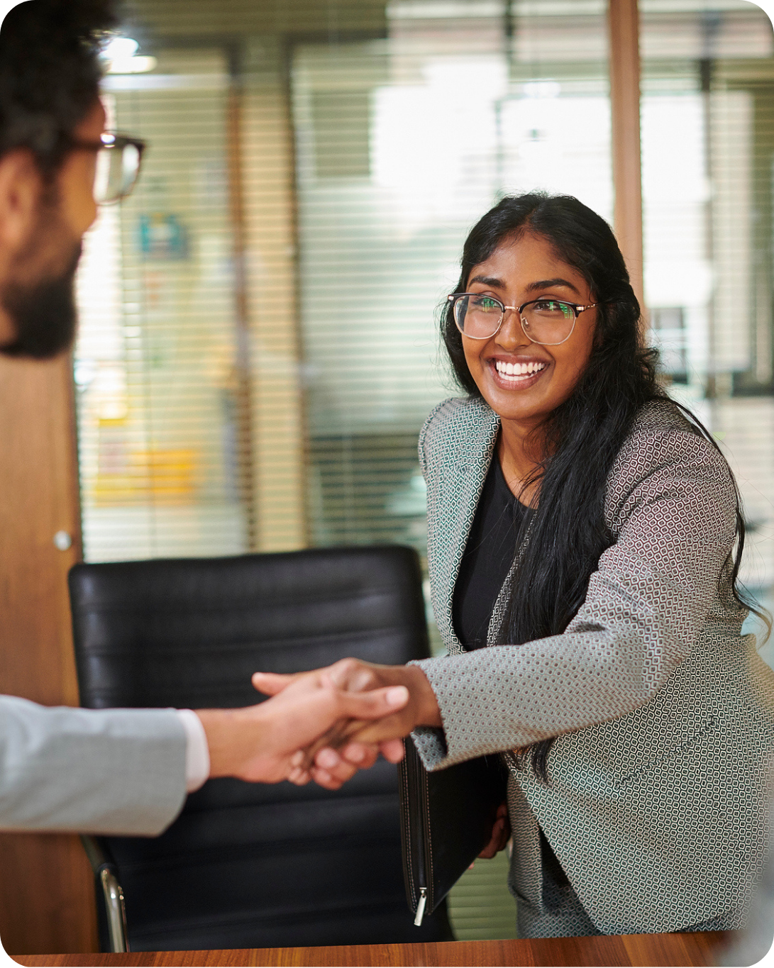 A woman in business attire smiles and shakes hands with another person across a desk in an office setting.
