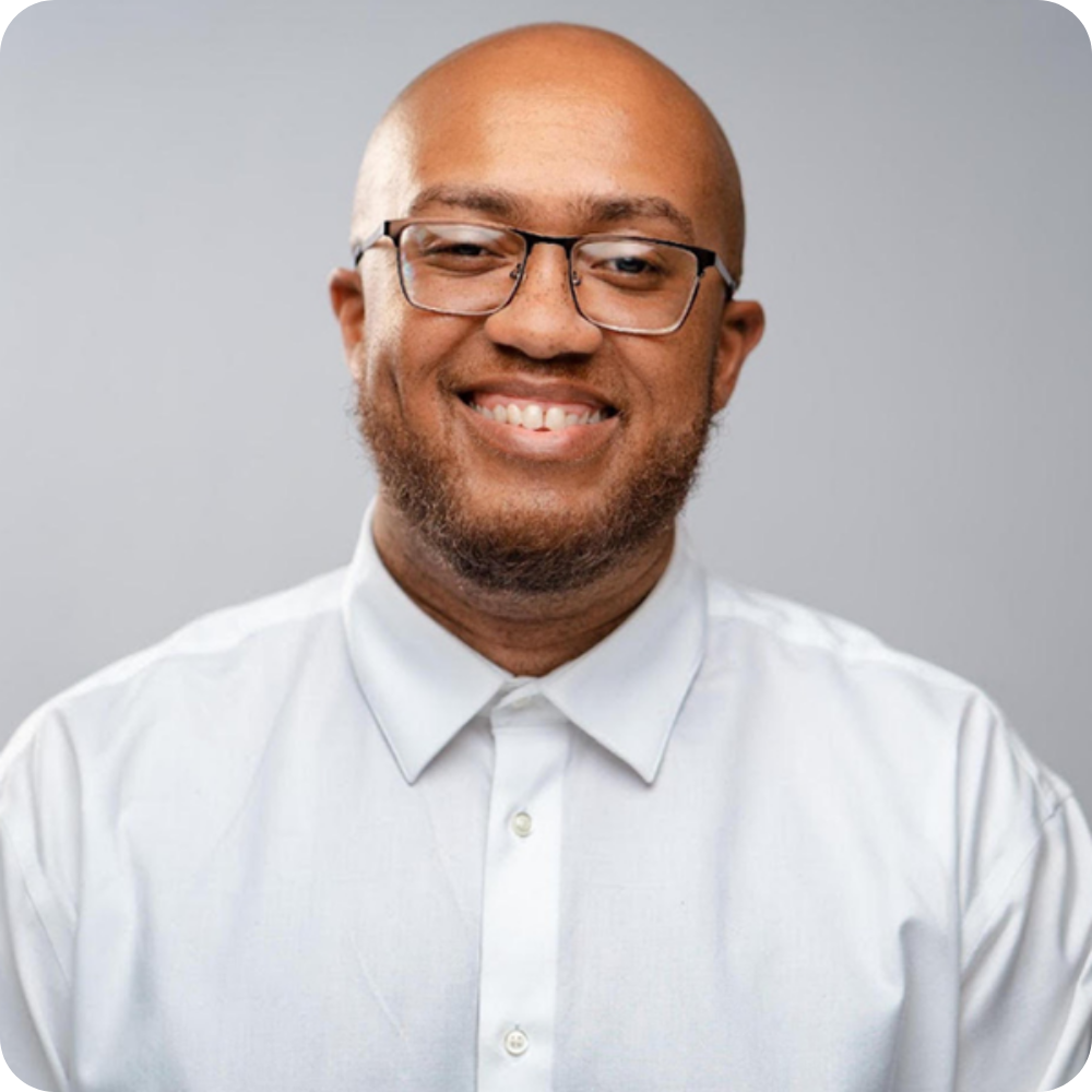 A man wearing glasses and a white button-up shirt smiles at the camera against a plain gray background.