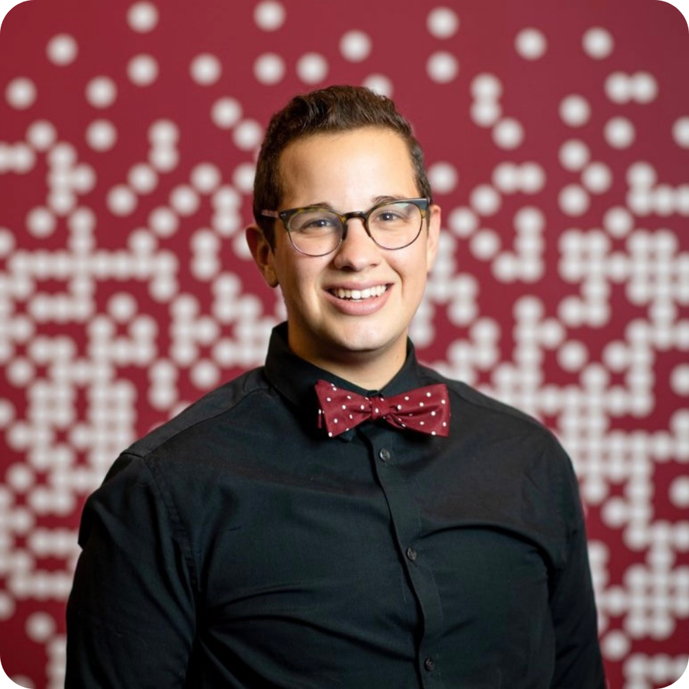 A person wearing glasses, a black shirt, and a red polka dot bow tie stands smiling in front of a red background with a white dot pattern.