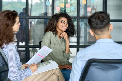 Three people sit in an office having a meeting; one woman smiles while another holds documents, and colorful sticky notes are posted on the glass wall behind them.