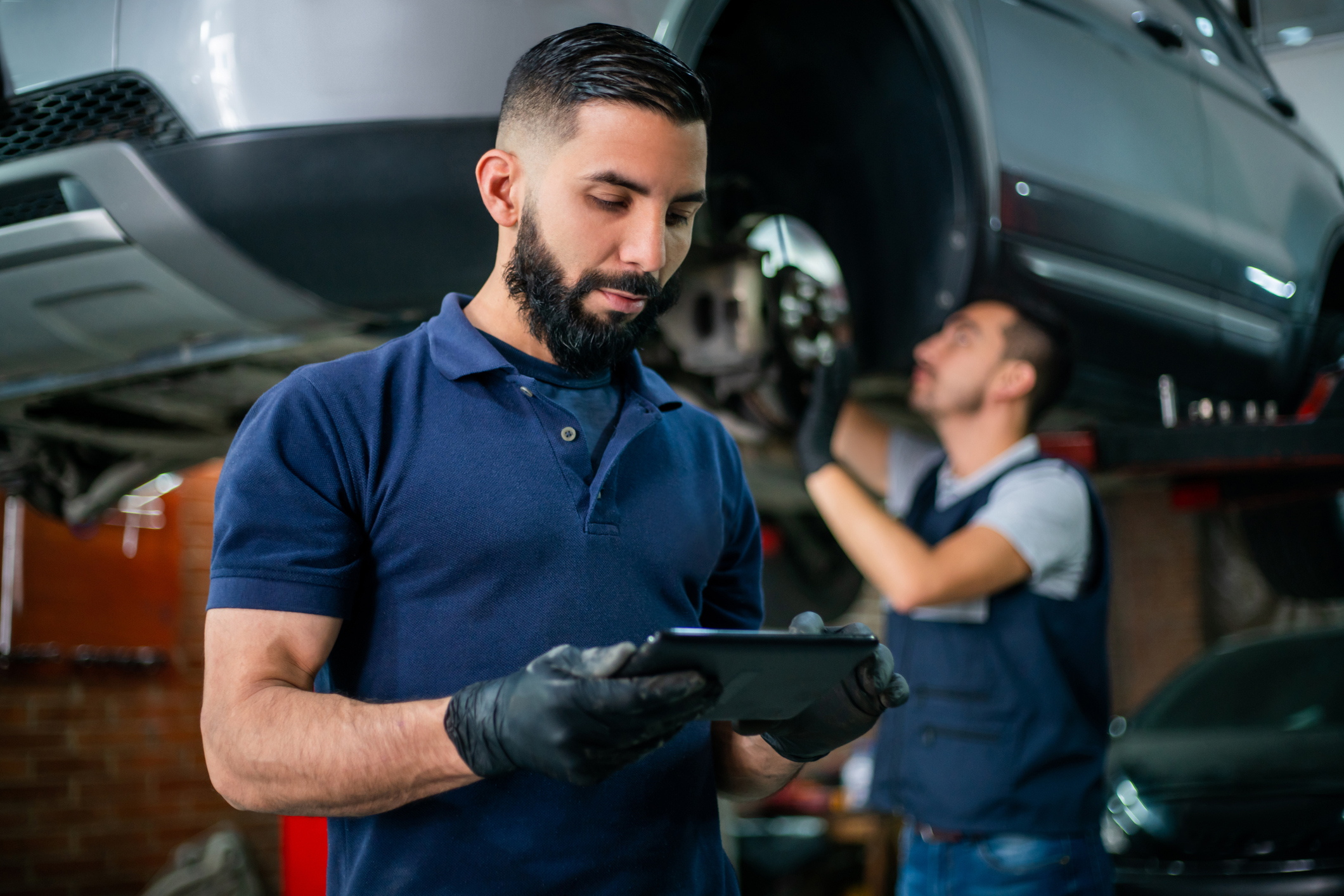 A mechanic wearing gloves uses a tablet while another mechanic works on a car lifted in a garage.