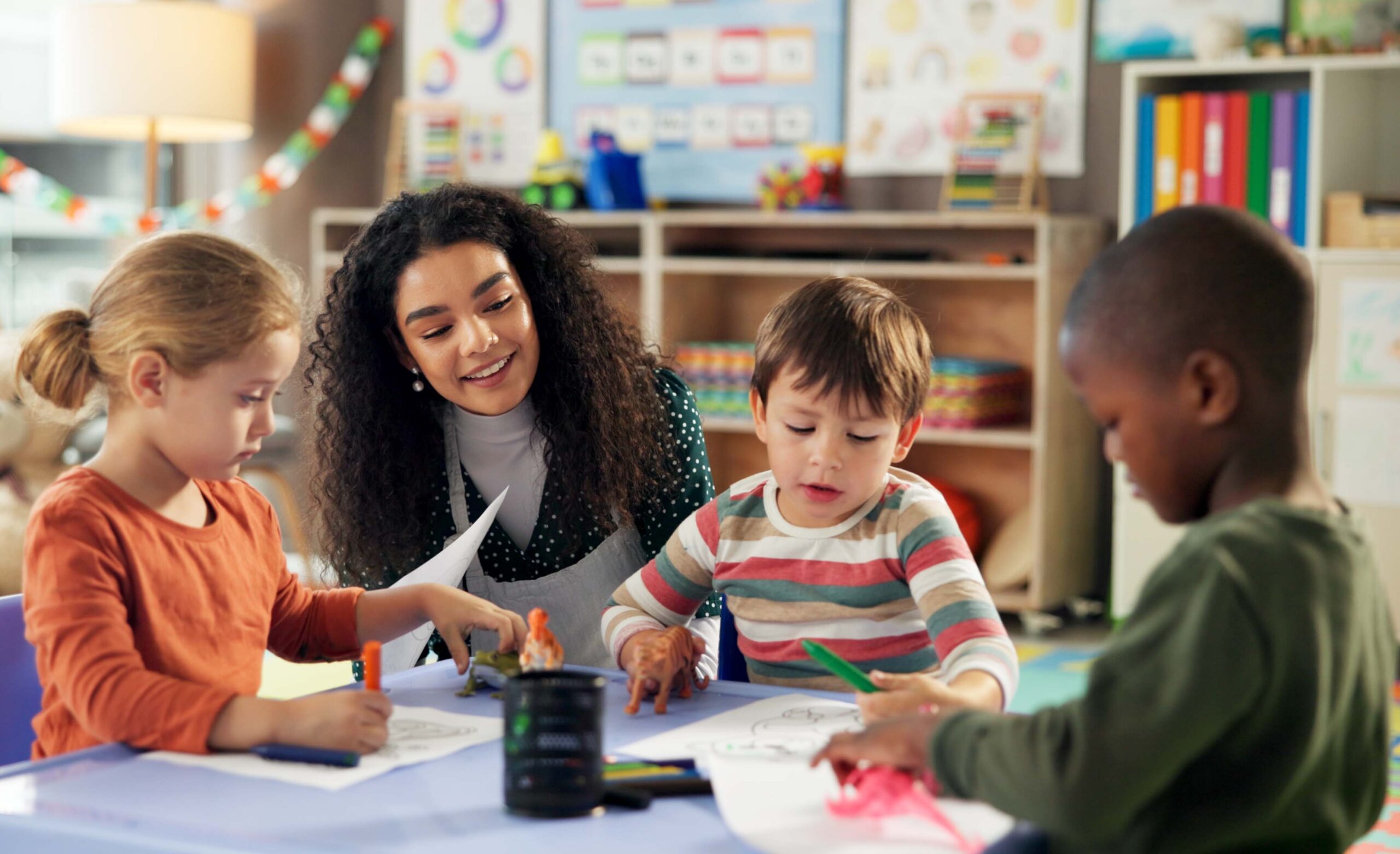 A teacher sits at a table with three young children as they draw with crayons and markers in a classroom setting.
