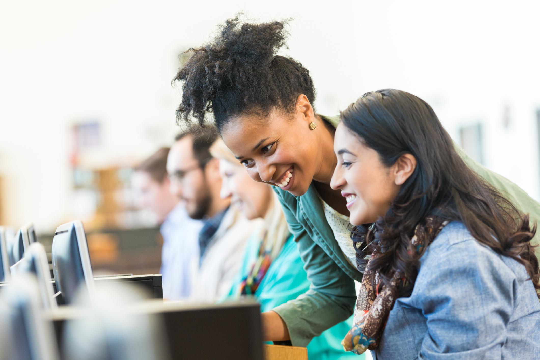 Two women smile and work together at a row of computers in a bright room, with other people using computers in the background.